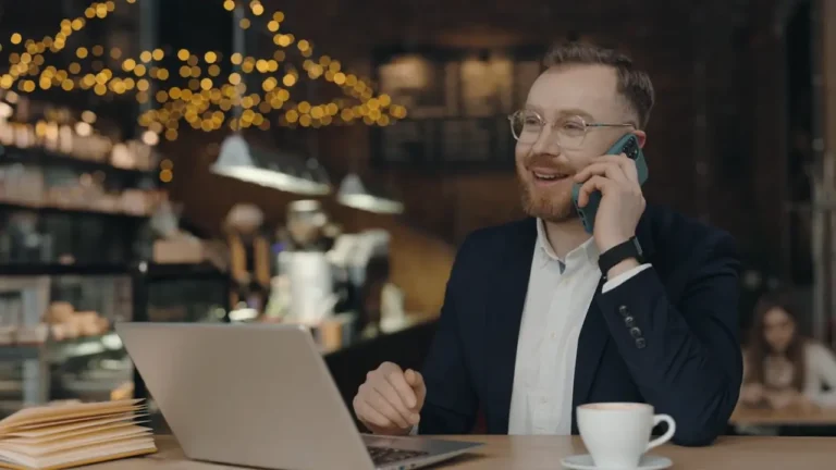 Man in casual suit, talking on his cell phone in a restaurant. A laptop and coffee cup are in front of him.